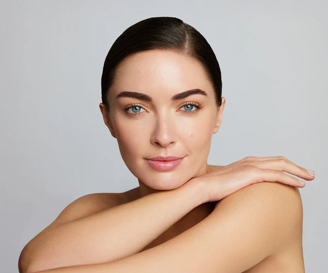 Portrait of a woman with sleek dark hair, glowing skin and natural makeup, resting her arms across her shoulder against a light grey background.