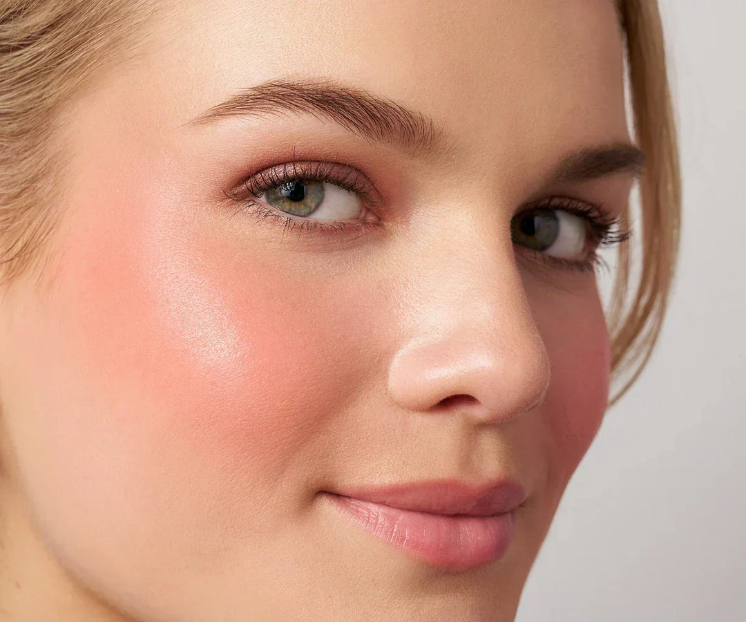 Close up model photo showing bright and bold peachy pink liquid blush applied to the cheeks. Complexion is clean and fresh.
