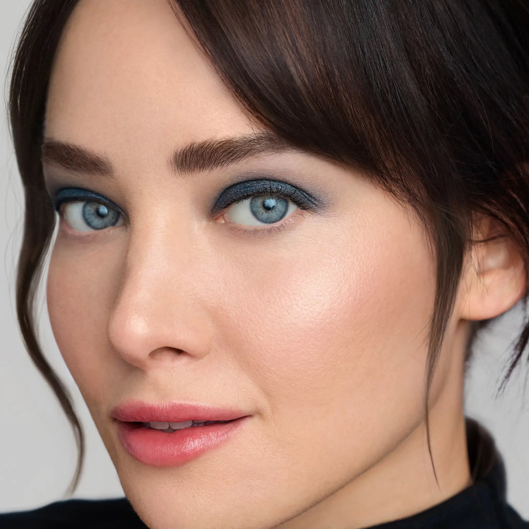 Model wearing Jane Iredale® ColorLuxe Eye Shadow Stick in Riviera, a deep shimmering blue, paired with Jane Iredale® PureBronze Shimmer Bronzer in First Light, glowing skin and a soft pink lip.