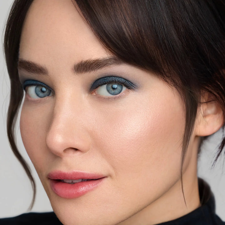 Model wearing Jane Iredale® ColorLuxe Eye Shadow Stick in Riviera, a deep shimmering blue, paired with Jane Iredale® PureBronze Shimmer Bronzer in First Light, glowing skin and a soft pink lip.