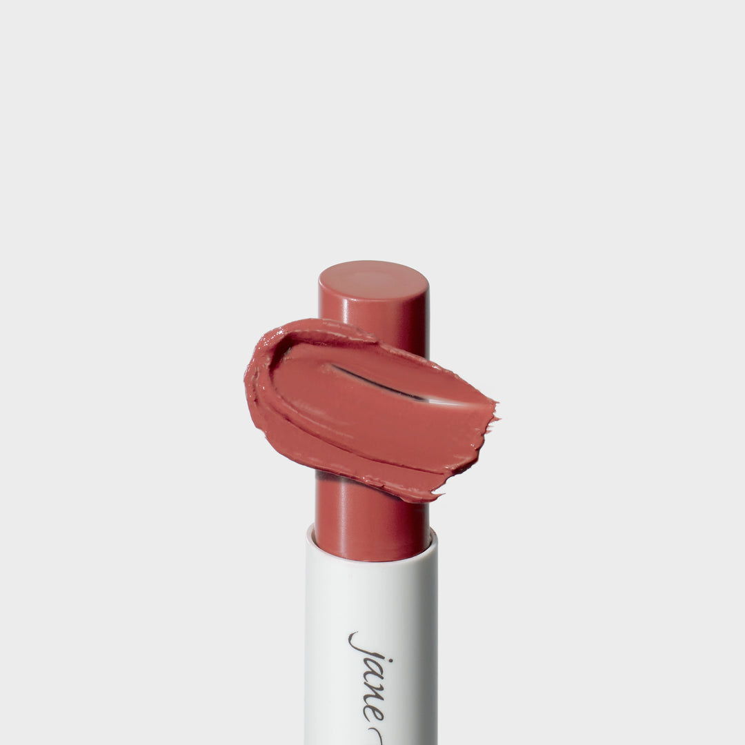 Jane Iredale® lipstick in Rosebud, shown in a white tube with the lipstick bullet extended and a warm rosy nude cream smear sweeping across the front.