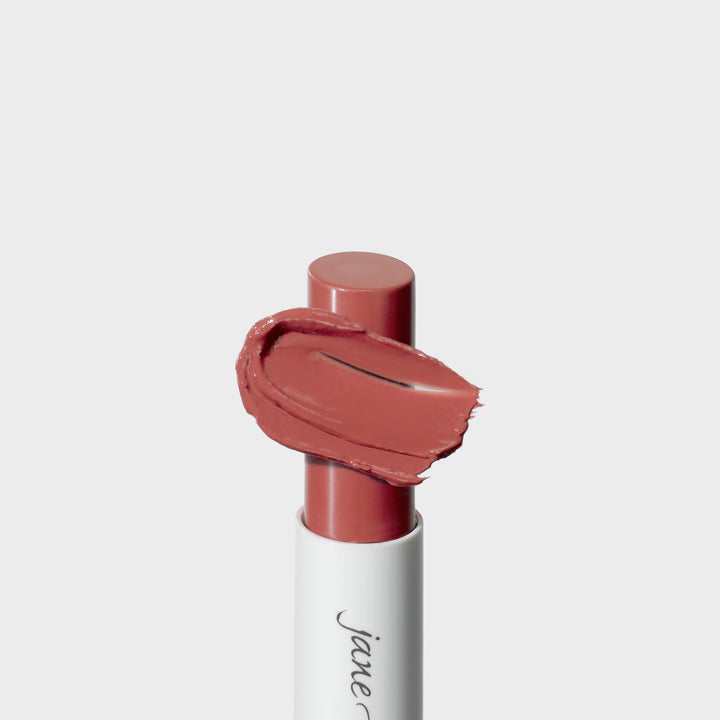 Jane Iredale® lipstick in Rosebud, shown in a white tube with the lipstick bullet extended and a warm rosy nude cream smear sweeping across the front.