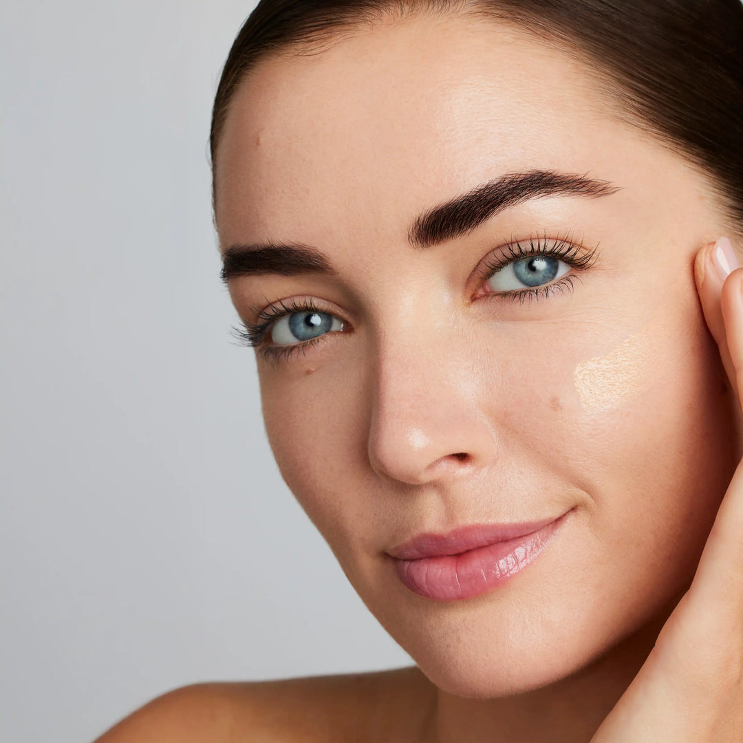 Close-up of a woman's face with a neutral background. Model has a swipe of foundation across her cheek that matches the natural colour of her complexion
