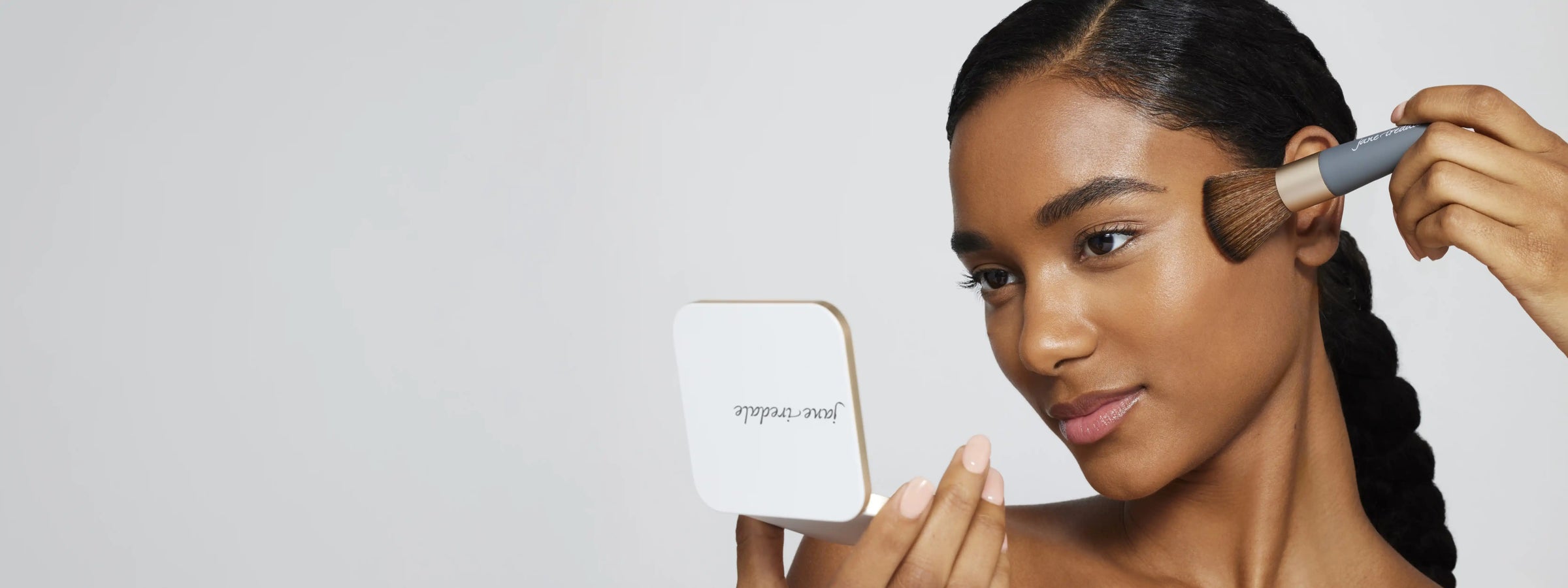 Jane Iredale model holding a refillable compact and looking into the mirror as she applies powder foundation with the Handi Brush