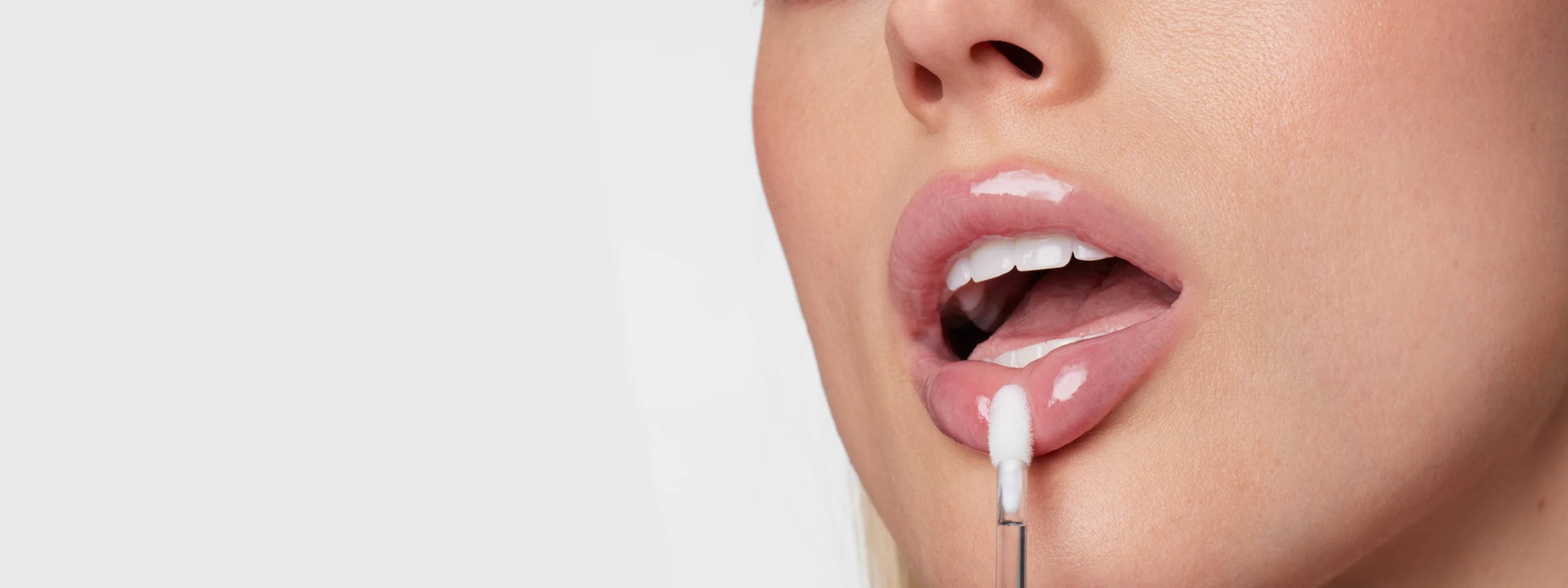 Close-up of a model applying glossy pink lip product to the lower lip with a soft doe-foot applicator, showcasing luminous skin, defined lashes and a fresh, polished beauty look.